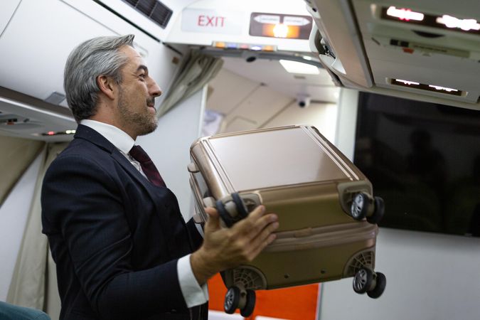 Male in business attire putting small suitcase above seats in airplane cabin