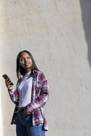 Female standing in the sun in front of wall holding phone and looking around, vertical