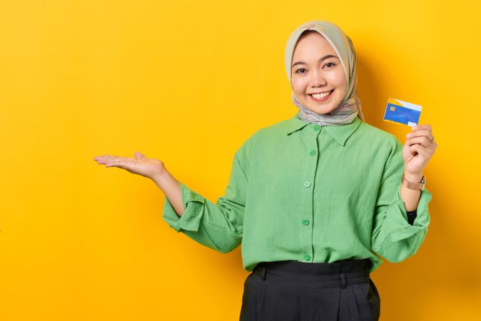 Muslim woman in headscarf and green blouse holding credit card with palm up on copy space