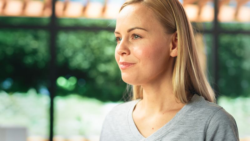 Authentic Portrait of Young Female in Kitchen Room at Home Looking Thoughtfully at Her Backyard. Attractive Blond Woman Poses for Camera and Smiles.