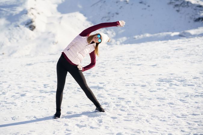 Female in hat, polarized sunglasses, stretching sideways on on snowy mountain