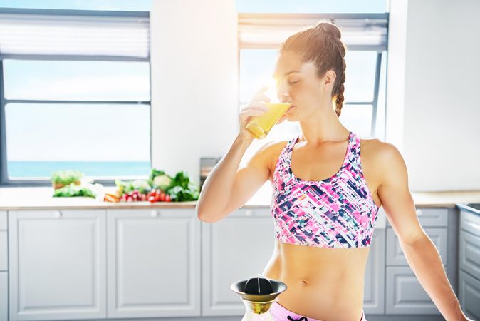 Muscular woman sipping fresh orange juice in bright kitchen