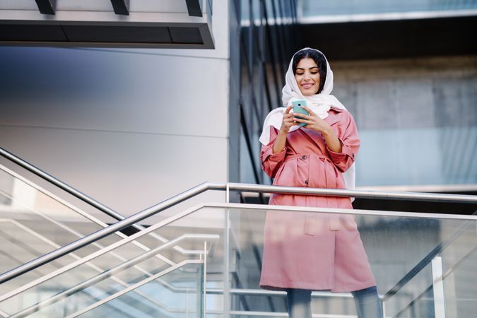 Female in headscarf and pink trench coat standing on staircase with phone