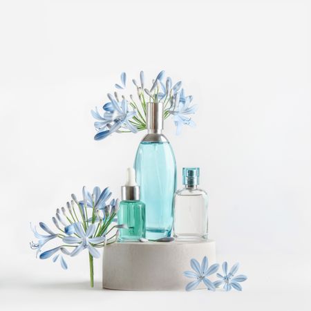 Beauty products in blue glass spray and pipette bottles standing on podium with flowers and petals at white background. Cosmetic setting with serum,perfume fragrance and toner. Front view