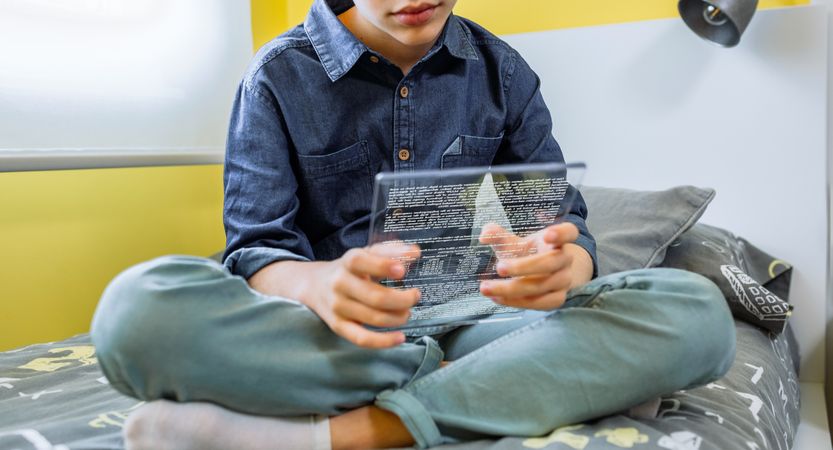 Unrecognizable boy using futuristic transparent tablet to read ebook sitting over his bed