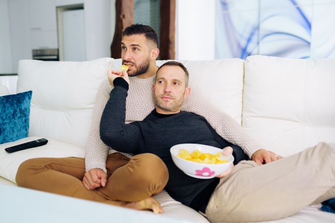 Two men relaxing on sofa together with bowl of chips