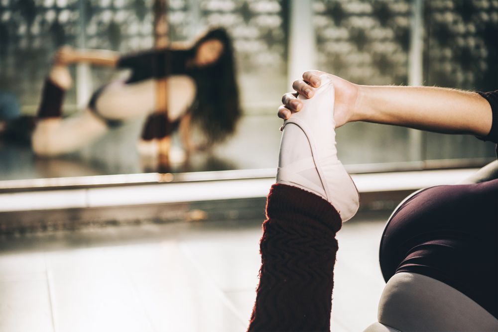Crop shot of ballerina pulling leg to stretch muscles sitting on floor in ballet studio.