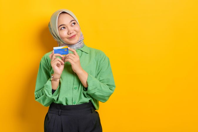 Muslim woman in headscarf and green blouse holding credit card with both hands looking away