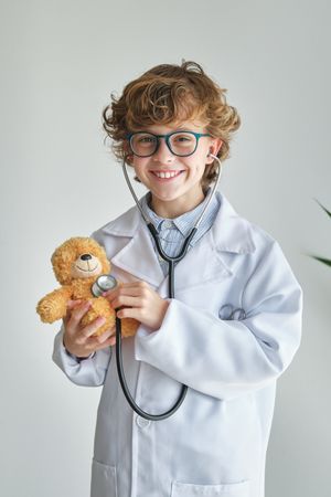Cheerful boy in medical uniform examining toy bear with stethosc