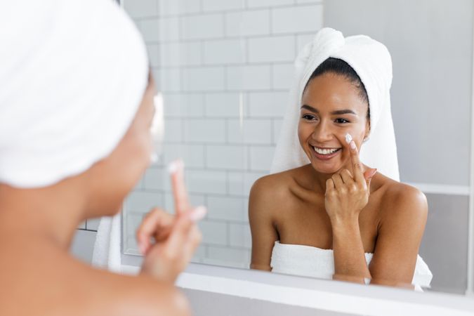 Black woman in towel applying face cream in the mirror