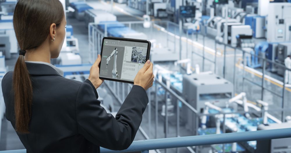 Thoughtful Female Engineer Monitoring and Analyzing Conditions at a Modern Tech Electronics Factory with Automated Robot Arms Working with the Help of Tablet Computer