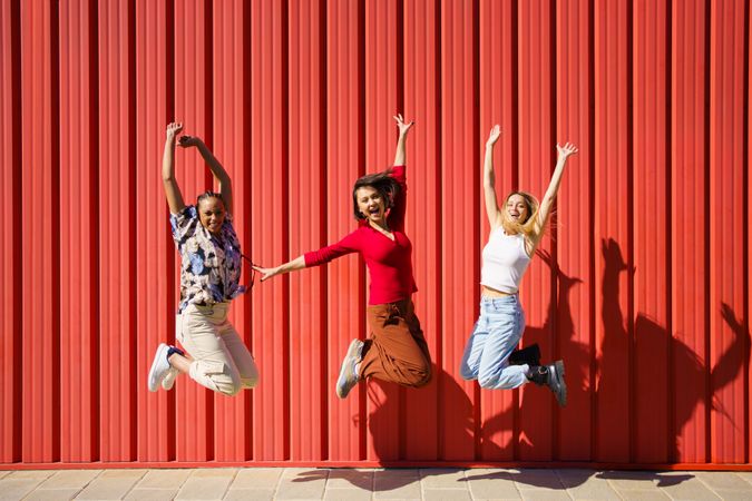 Three females jumping for joy in front of red wall on sunny day