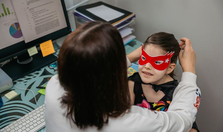 Woman teleworking and putting costume mask on her daughter