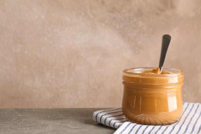 Glass jar with peanut butter and spoon, and kitchen towel on wooden table against light background, space for text