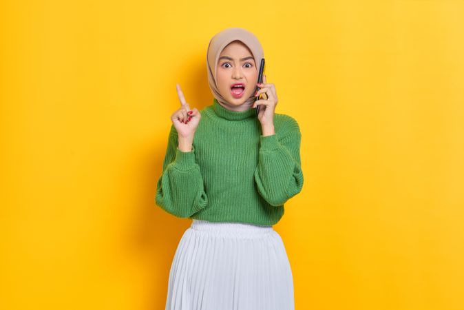 Surprised woman in headscarf holding up finger while talking on phone