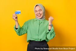 Muslim woman in headscarf and green blouse holding credit card and clenching fist with eyes closed 4OAvZ0