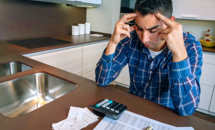 Stressed man with calculator and bills in kitchen