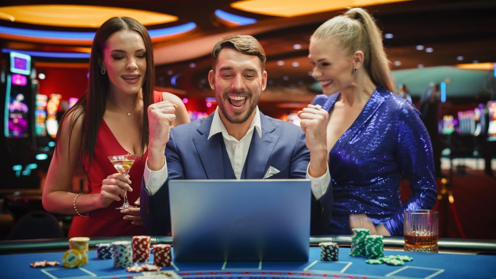 Portrait of a Happy Man with Two Female Partners Betting Online on a Laptop Computer while Sitting in a Luxury Casino.