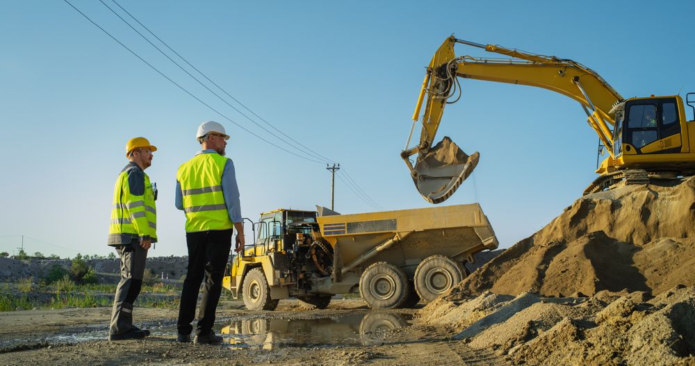 Male Real Estate Investor And Project Manager Talking On Construction Site Of Apartment Block. Colleagues Discussing Progress, Business. Excavator Loading Sand In Industrial Truck.