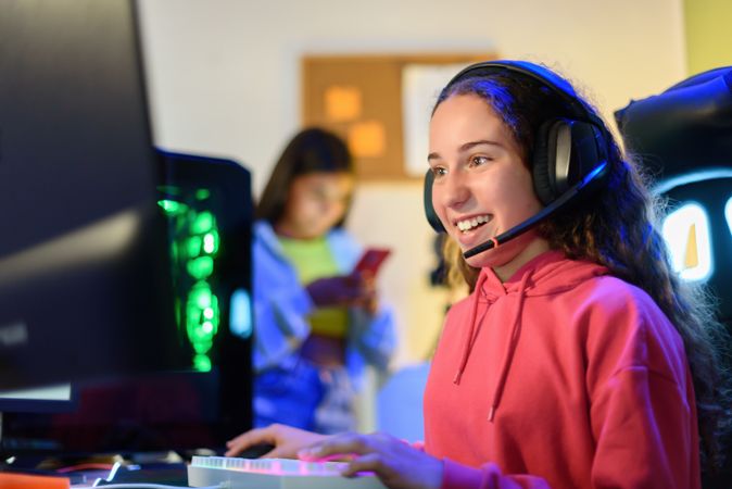 Happy teenage girl in headphones sitting at table and playing games in room