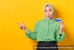 Shocked Muslim woman in headscarf and green blouse holding credit card and pointing aside 0geJj4