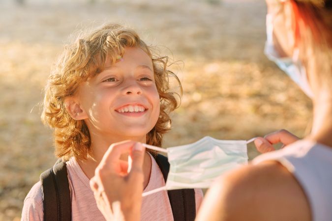 Unrecognizable woman putting mask on cheerful boy