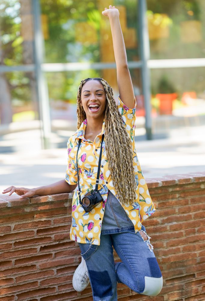Smiling female in bold patterned shirt leaning on brick wall with camera, vertical