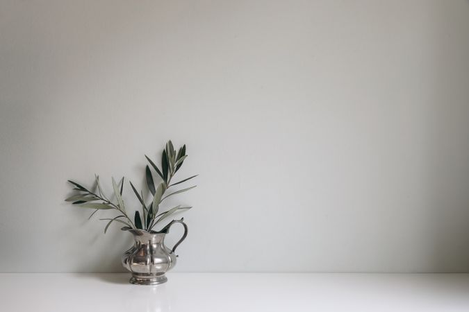 Olive tree branches, twigs in silver jug vase on table. Empty mint green wall mockup, background. Working space, home office decor. Vintage Mediterranean summer interior still life.