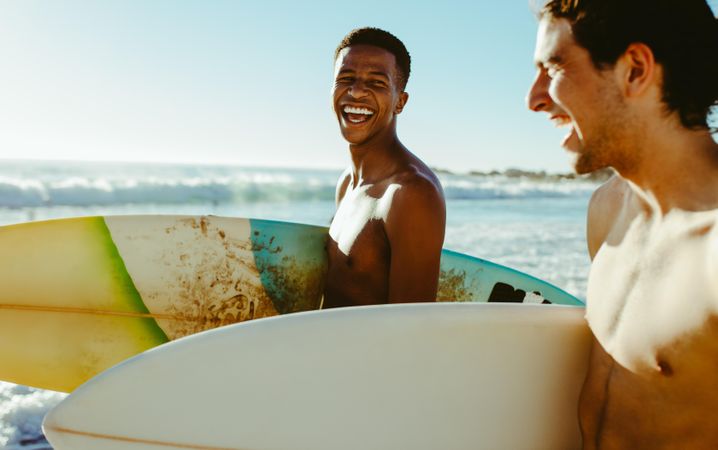 Happy young friends walking on beach carrying surfboards
