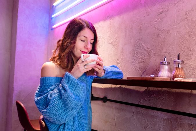 Positive woman with cup at table near wall with neon lamps