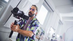 Focused Young Male Videographer With Beard, Filming In A Bright Studio, Holding A Professional Camera, Surrounded By Modern Filmmaking Equipment, Engaged In A Creative Production Process.