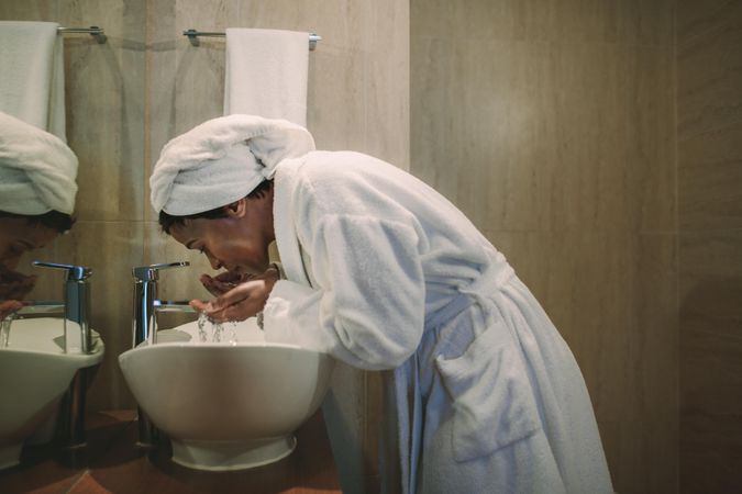 Woman in bathrobe washing her face in bathroom sink