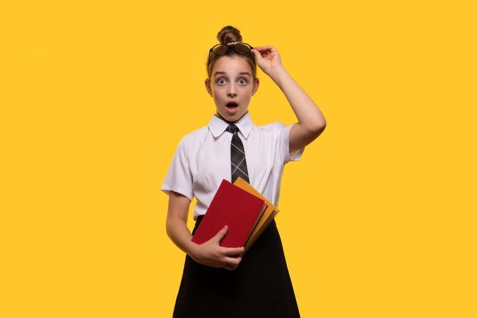 Surprised teen girl holding books, wearing school uniform, lifting glasses.