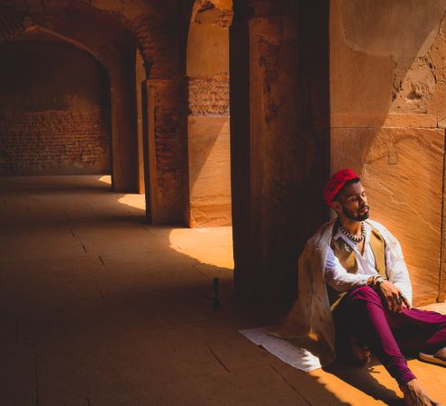 Man in traditional outfit sitting on floor leaning on pillar