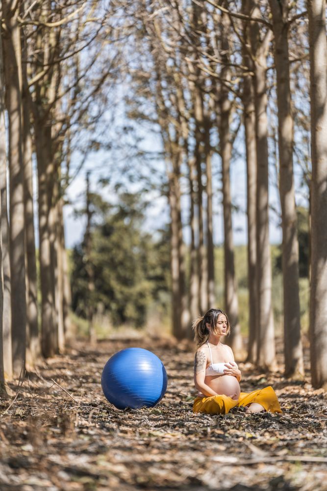 Serene adult pregnant woman meditating while sitting on ground among trees in park during sunny day