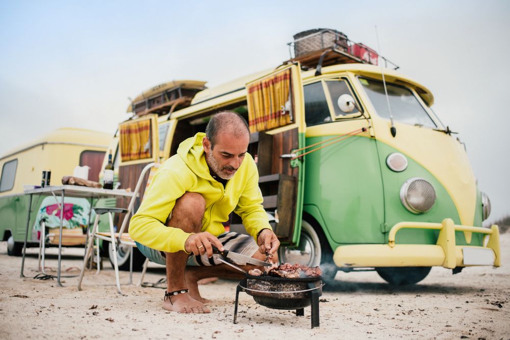 Tanned man cooking on camping stove beside parked mini van