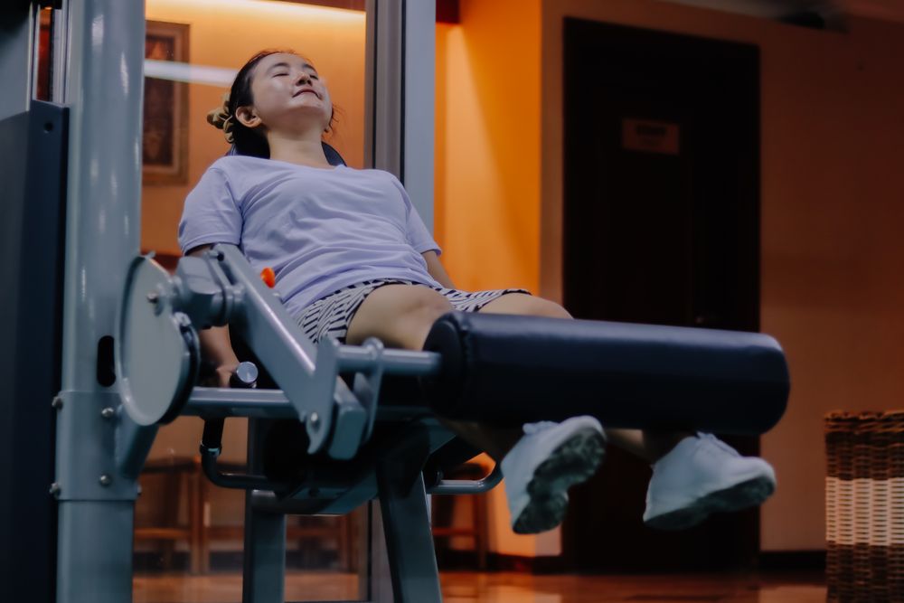 woman wearing purple shirt, shorts, and white shoes, is using a leg extension exercise machine.