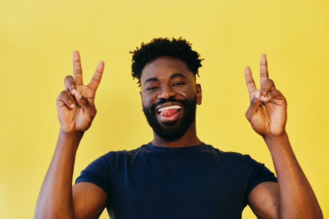 Black man in navy t-shirt in yellow studio making the peace sign with both hands