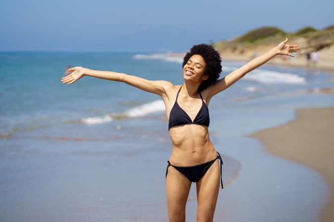 Woman in bikini standing on beach with outstretched arms and eyes closed