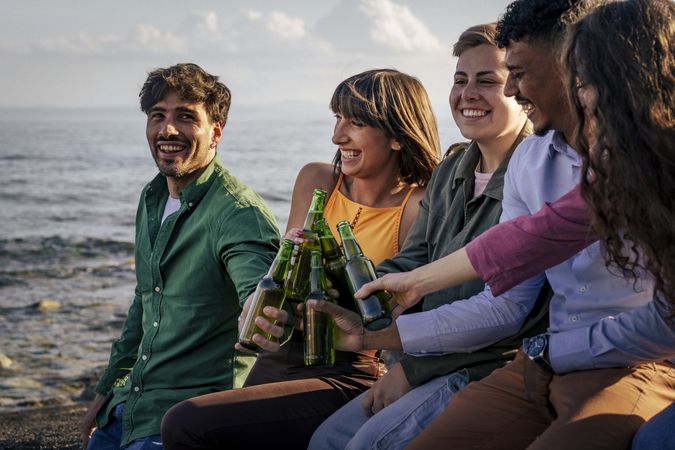 Group of diverse friends laughing and toasting with beer on the beach at sunset