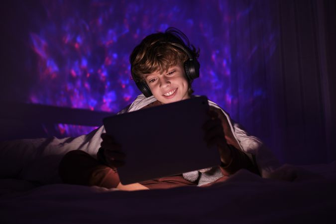 Positive boy in headphones watching video on tablet in darkness
