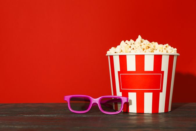 3d glasses and striped bucket with popcorn against red background