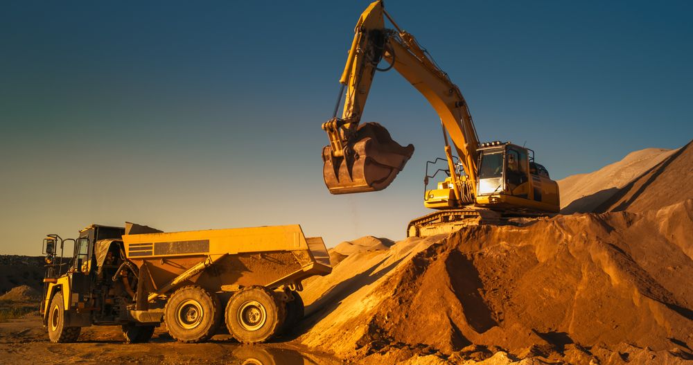 Construction Site On Sunny Evening: Industrial Excavator Loading Sand Into A Truck. The Process Of Building New Apartment Block. Workers Operating Heavy Machinery To Complete A Real Estate Project.