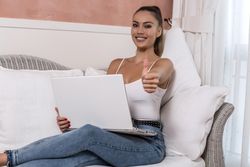 Smiling woman using computer with thumb up sitting on couch