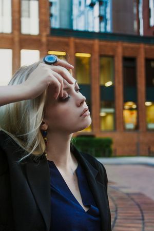 Blonde woman in dark blazer touching her forehead standing outdoor