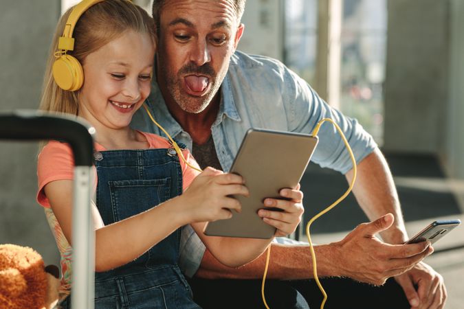 Cute girl wearing headphones looking at digital tablet with her father sticking out tongue