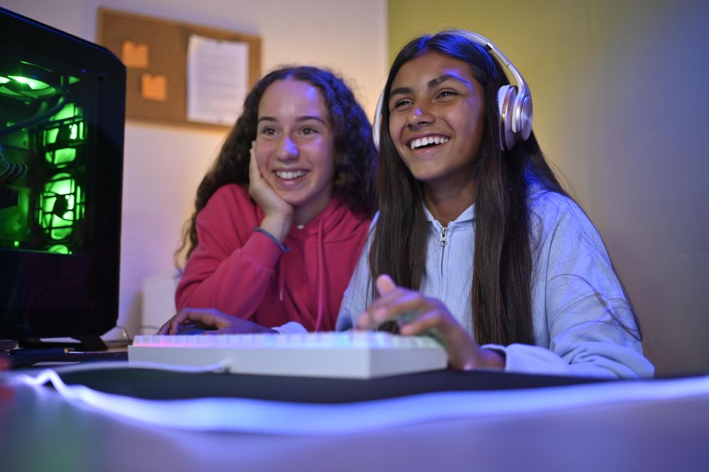 Cheerful teenage diverse girlfriends sitting at table with gaming PC and playing game