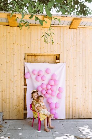 Girl embracing toddler on chair against wall with pink balloons