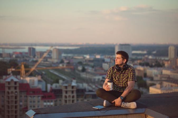 Male with tablet, headphones and takeaway coffee cup while enjoying view from rooftop