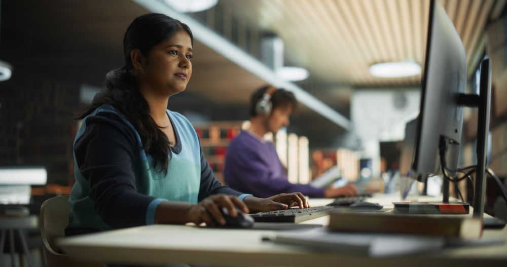 Thoughtful Indian Female Student Working on Personal Development in a Quiet Public Library with Rows of Bookshelves. Young Smart South Asian Woman Preparing for Examination in College